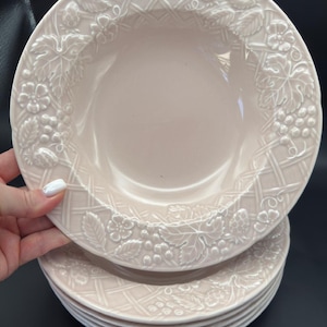 May include: A stack of pale pink ceramic bowls. Each bowl has a raised, decorative border with a lattice pattern and embossed floral and fruit motifs, including strawberries and grapes. The bowls have a shallow, rounded shape.