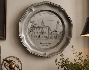 May include: A silver-colored decorative plate with a scalloped edge. The plate features a detailed black and white illustration of a building with a steeple and the text "Hemau/Opf. Rathaus". The plate is hanging on a light-colored wall.