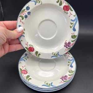 Puede incluir: Una pila de platillos de cerámica blanca con un borde decorativo de frutas y flores. El borde presenta manzanas rojas, uvas moradas y bayas azules con hojas verdes y detalles azules. Los platillos tienen un área central empotrada para una taza.