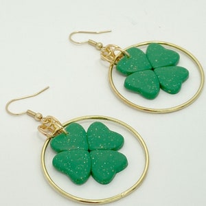May include: A pair of gold hoop earrings with green four-leaf clover charms. The earrings are handmade and feature the text "HER BLOOM" and "@herbloom".