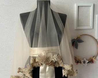 May include: A sheer ivory veil with a gold beaded trim and a matching ivory satin sash. The veil is draped over a mannequin.