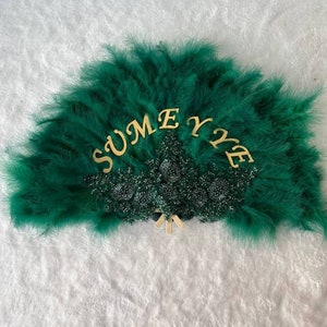 Puede incluir: Un abanico de plumas verdes con letras doradas que dicen "SUMEYE". El abanico está decorado con cuentas verdes.