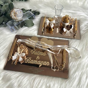 Peut inclure: Deux plateaux miroir avec des accents dorés et des éléments décoratifs. Le plateau supérieur présente une tasse en verre, une boîte dorée avec un ruban blanc et une boîte dorée avec un ruban blanc et un pompon doré. Le plateau inférieur présente deux pièces en forme de cœur avec les noms "Fatma & Ramazan" et un ruban blanc avec le texte "Fatma Ramazan".