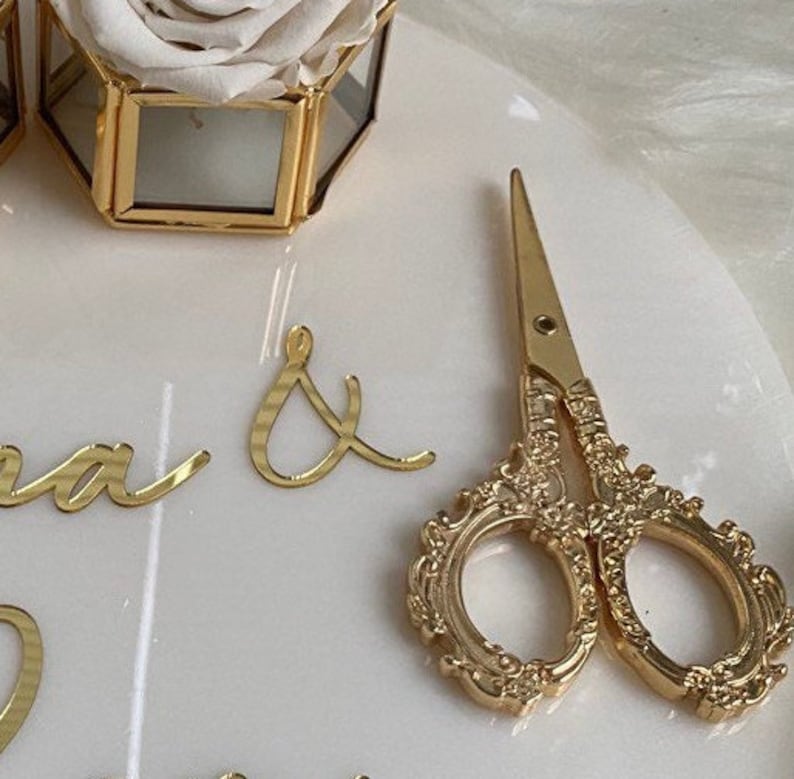 Op de afbeelding: Gouden schaar met een sierlijk ontwerp op een wit oppervlak. De schaar is open en de messen zijn scherp. Het oppervlak is glad en heeft een lichte glans.