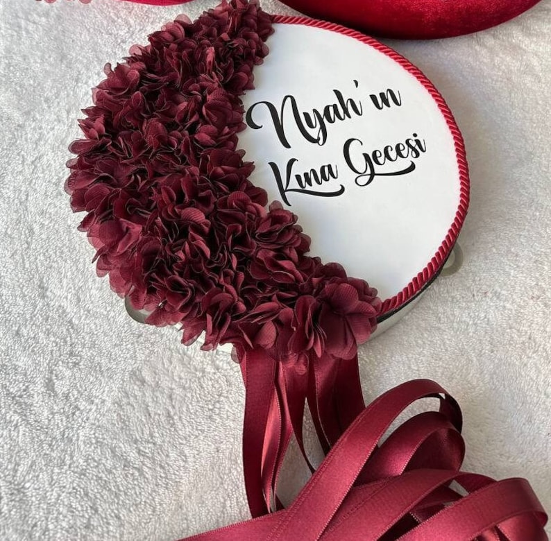 May include: A white drum with a red trim and a maroon fabric flower design. The drum has the text "Nyah'in Kına Gecesi" written in black.