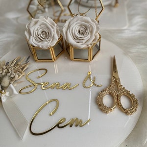 Op de afbeelding: Een wit rond bord met gouden letters die "Sena & Cem" zeggen. Twee zeshoekige glazen dozen met gouden randen bevatten witte rozen. Een paar gouden scharen ligt op het bord.