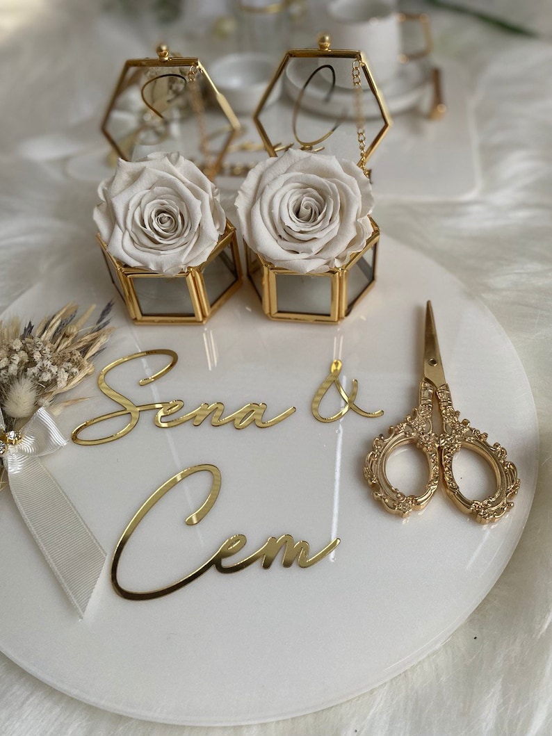 Op de afbeelding: Een wit rond bord met gouden letters die "Sena & Cem" zeggen. Twee zeshoekige glazen dozen met gouden randen bevatten witte rozen. Een paar gouden scharen ligt op het bord.