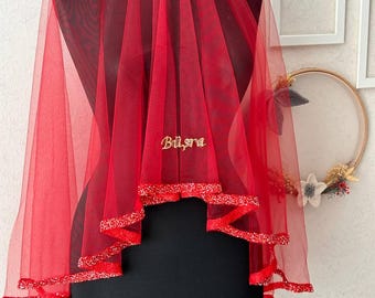 May include: A red tulle veil with a gold embroidered name "Bilsa" on the front. The veil has a beaded trim around the edges.