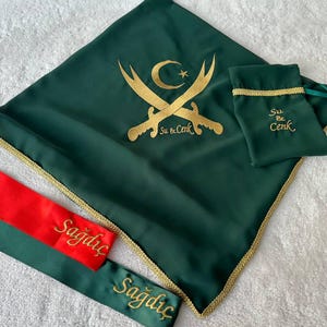 May include: A green fabric square with gold trim and a gold embroidered design of crossed swords and a crescent moon with a star. The text "Su & Cenk" is embroidered below the design. A smaller green fabric square with gold trim and the text "Su & Cenk" is in the upper right corner. Two green fabric sashes with gold trim and the text "Sağdıç" are in the lower left corner.