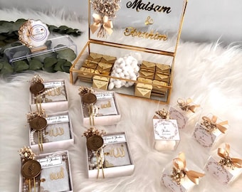 May include: An assortment of wedding favors and gifts, including a gold-framed glass box with chocolates, small gift boxes with floral accents, and small books with decorative elements. The favors are arranged on a white, fluffy surface.