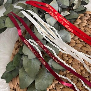 May include: Close-up of personalized ribbons in red, white, and burgundy. The ribbons are adorned with gold text and paired with strings of pearls. Eucalyptus leaves and a woven basket provide a natural backdrop.