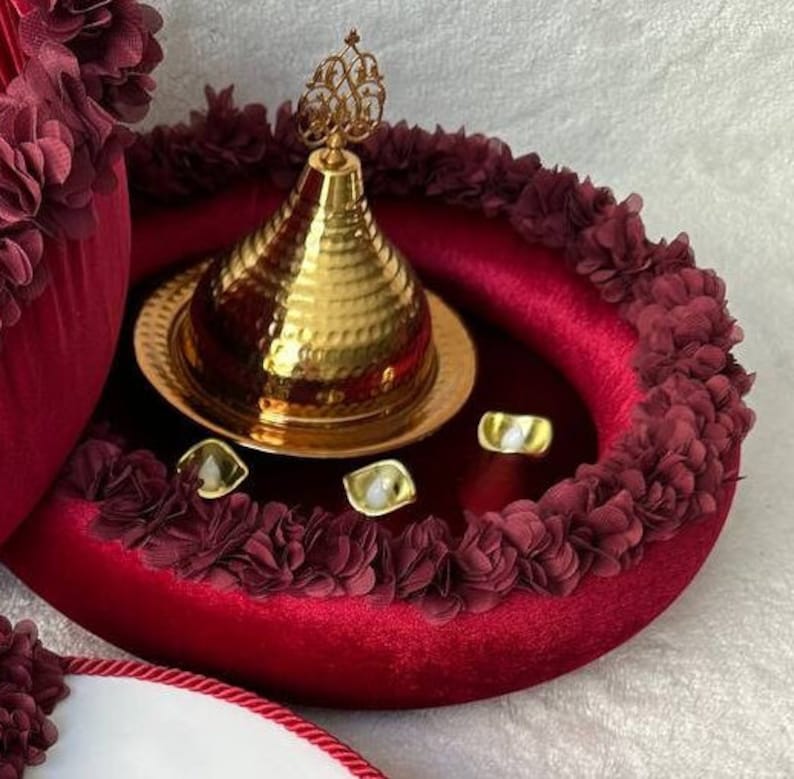 May include: A red velvet tray with a gold decorative lid and a gold base. The tray is decorated with burgundy fabric flowers around the edge. There are four small white decorative items on the tray.