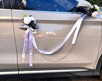 Può includere: Un'auto colorata di chiaro decorata per un matrimonio. Composizioni floreali bianche e lunghi nastri bianchi adornano le maniglie delle porte e lo specchietto laterale. I nastri hanno del testo stampato sopra. Una catena collega i nastri.
