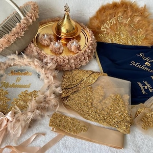 Pode incluir: Um conjunto de cerimónia de casamento com decorações douradas e bege. O conjunto inclui uma bandeja com um bule dourado, uma cesta com flores bege, um prato decorativo com glitter dourado, um prato decorativo com penas, um prato decorativo com glitter dourado e um tecido azul com os nomes "Suheila & Mahmud" bordados.
