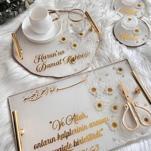 May include: A white tray with gold accents and the text "Harun's Groom's Coffee" in gold lettering. The tray is decorated with white flowers and pearls. A white coffee cup and saucer, a clear glass, and a pair of gold scissors are on the tray. A second white tray with gold accents features a gold Arabic inscription and the text "And Allah, joined their hearts with love.  Anfal 8/63".