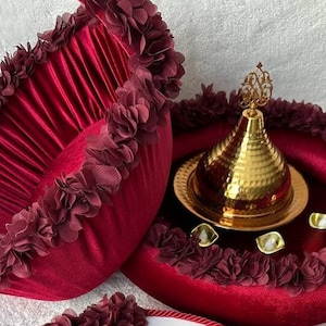 May include: A burgundy velvet tray with a gold metal lid and decorative trim. The tray is surrounded by a burgundy velvet border with a ruffled edge.