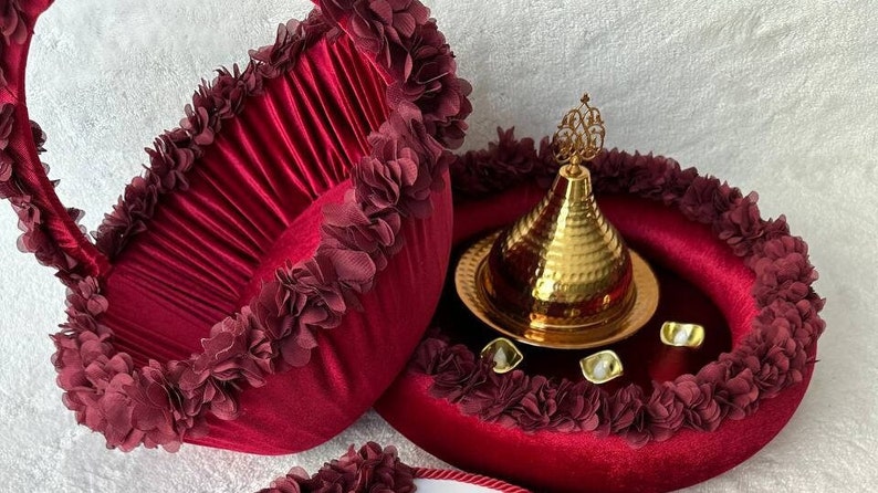 May include: A burgundy velvet tray with a gold metal lid and decorative trim. The tray is surrounded by a burgundy velvet border with a ruffled edge.