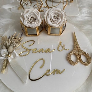 Op de afbeelding: Een wit marmeren dienblad met gouden accenten heeft twee glazen dozen met witte rozen, een klein boeket gedroogde bloemen en een gouden schaar. Het dienblad heeft ook de namen "Sena & Cem" in gouden letters.