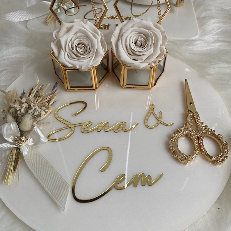 Op de afbeelding: Een wit marmeren dienblad met gouden accenten heeft twee glazen dozen met witte rozen, een klein boeket gedroogde bloemen en een gouden schaar. Het dienblad heeft ook de namen "Sena & Cem" in gouden letters.