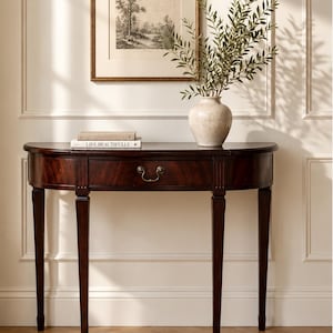 May include: A dark wood semi-circular console table with a single central drawer featuring a brass pull. On the table sits a beige ceramic vase with green foliage and a book with the text LIVE BEAUTIFULLY on the spine, below a framed print.