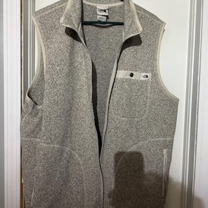 May include: A gray fleece vest with a full-length zipper and a stand-up collar. The vest has two side pockets and a chest pocket with a small logo. The vest is hanging on a green hanger against a white door.