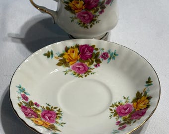 May include: A white teacup and saucer set with a floral pattern of pink and yellow roses. The set has a gold trim around the edges.