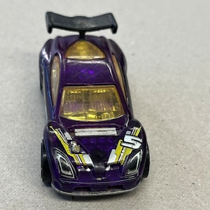 May include: Purple toy car with a black spoiler and yellow and white racing stripes. The car has a clear windshield and headlights.