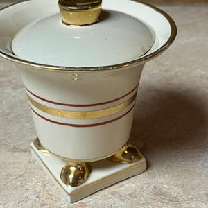 Puede incluir: Un tarro de cerámica blanco con tapa y base doradas. El tarro tiene dos rayas rojas con ribete dorado alrededor del centro. El tarro tiene cuatro patas doradas.