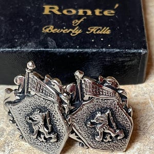 Puede incluir: Un par de gemelos de plata con un diseño heráldico con un león y una bandera. Los gemelos están en una caja negra con el texto "Ronte' of Beverly Hills".