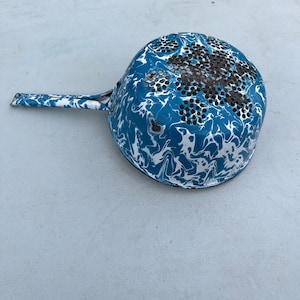 Puede incluir: Un colador de esmalte azul y blanco con un fondo de malla metálica negra. El colador tiene un patrón de mármol y un borde negro.