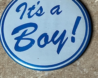 May include: A blue and white round button with the text "It's a Boy!" in blue cursive lettering.