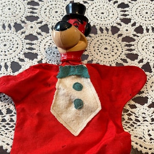 May include: A red felt hand puppet with a brown and black dog head wearing a black top hat. The puppet has a white felt bow tie with green felt accents.