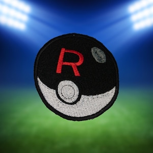 May include: A circular black patch with a red "R" and a white and gray design. The patch has a white border and a gray spot on the upper right side. The background is a blurred image of a green field and blue sky with stadium lights.
