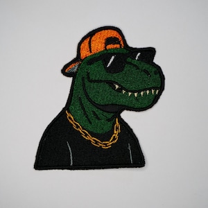 Puede incluir: Parche bordado con un Tyrannosaurus Rex verde que lleva gafas de sol, una gorra de béisbol naranja y gris y una cadena dorada. El dinosaurio tiene una expresión segura de sí mismo, sobre un fondo blanco.