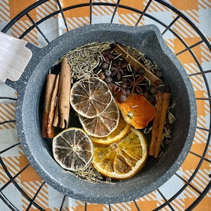 May include: A gray speckled pan filled with dried orange and lemon slices, cinnamon sticks, star anise, and rosemary. The pan sits inside a black metal trivet on an orange and white checkered surface. A wooden spatula rests on the pan's edge.