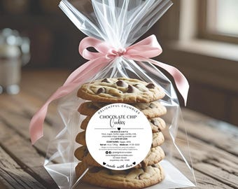 May include: A clear cellophane bag tied with a pink ribbon, containing six chocolate chip cookies. The cookies have a golden-brown color with visible chocolate chips. A round label on the bag reads "Chocolate Chip Cookies."