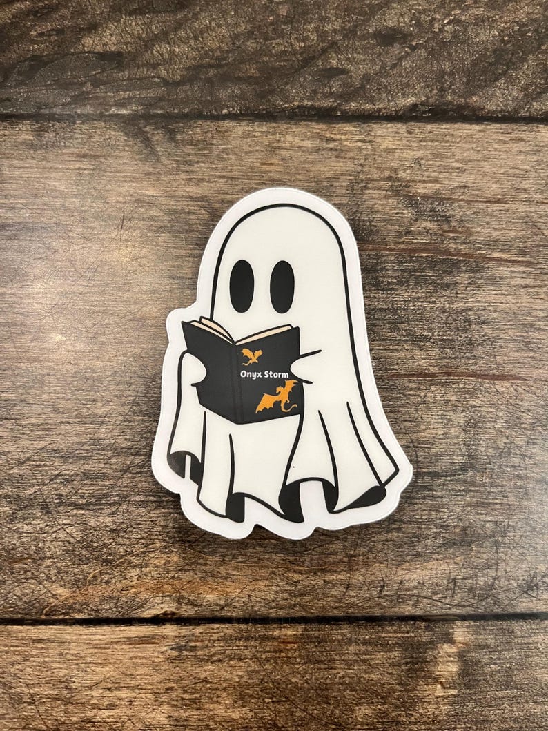 May include: A white ghost sticker with a black outline and black eyes. The ghost is holding a black book with the words "Onyx Storm" and an orange dragon illustration. The sticker is on a wooden surface.
