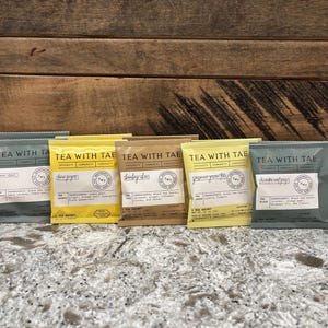 May include: Five tea sachets in various colors, including yellow, tan, and teal, are arranged in a row. Each sachet has the text "TEA WITH TAE" and a label with the tea type and ingredients. The sachets are on a speckled surface.