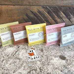 May include: Six colorful tea sachets from "Tea With Tae" are arranged on a countertop. The sachets are in shades of yellow, pink, brown, and green. A ghost sticker that says "I'm booooked" is in the foreground.