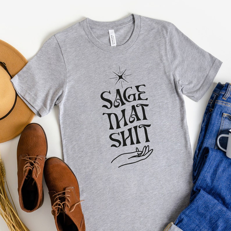 May include: Gray t-shirt with the text "Sage That Shit" in black lettering. A stylized hand with a star above it is also printed on the shirt.