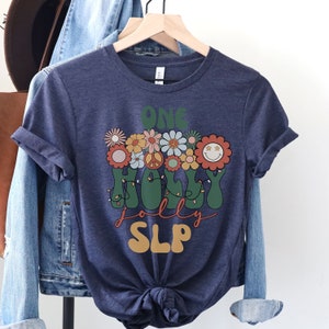 May include: A navy blue t-shirt with a retro-style graphic design featuring the text "One Holly Jolly SLP" surrounded by colorful flowers and peace signs.