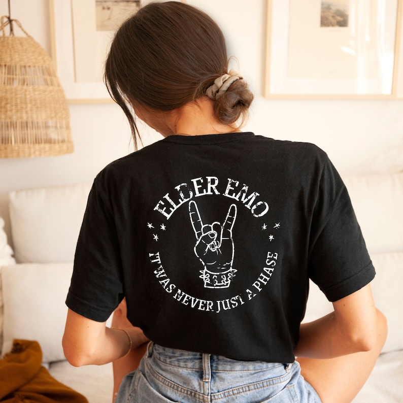 May include: Black t-shirt with a white graphic print of a hand making a rock and roll sign. The text "Elder Emo" is printed in a circle around the hand. The text "It was never just a phase" is printed below the hand.