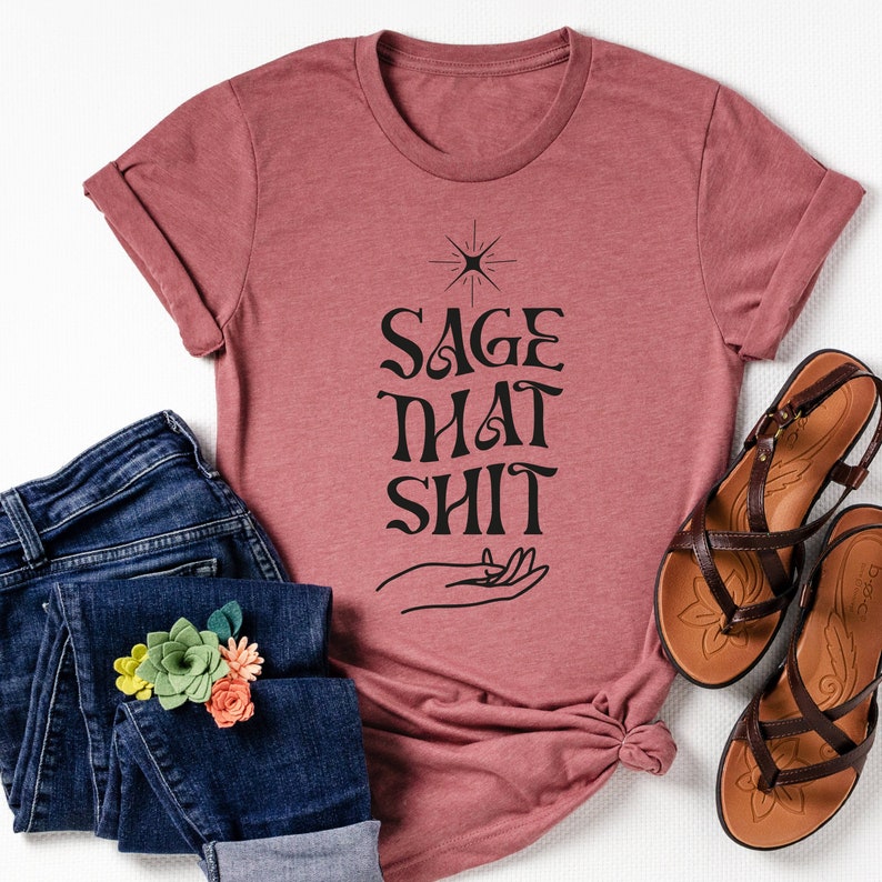 May include: A mauve t-shirt with a black graphic that says "Sage That Shit" with a hand holding a star above the text.