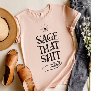 May include: A light pink t-shirt with a black graphic design that says "Sage That Shit" with a hand and a star design above the text.