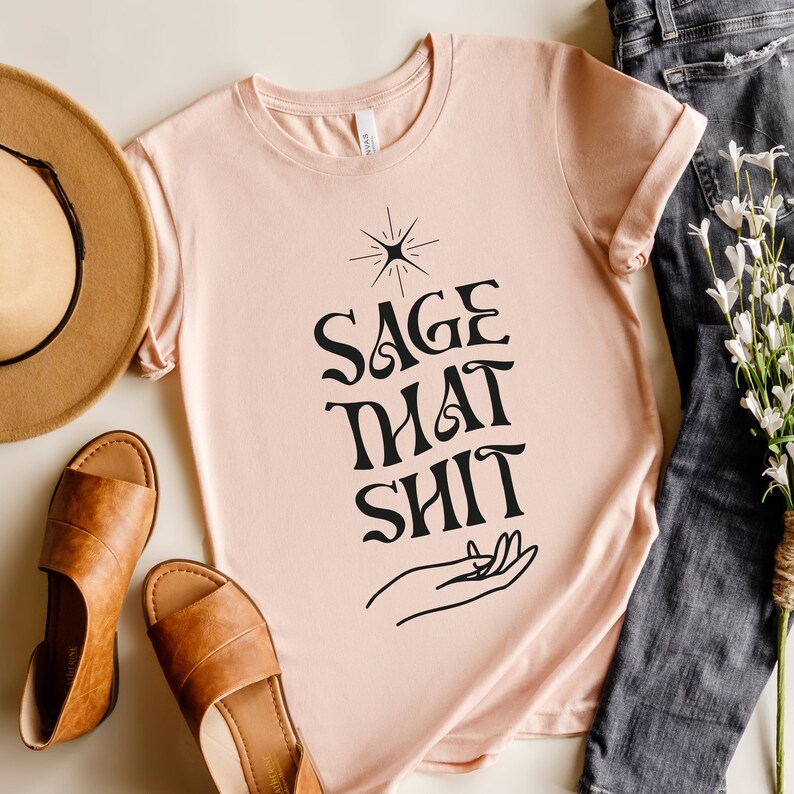 May include: A light pink t-shirt with a black graphic design that says "Sage That Shit" with a hand and a star design above the text.