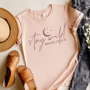May include: A pink t-shirt with the text "stay wild moon child" in a purple script font. A crescent moon and stars are above the text.