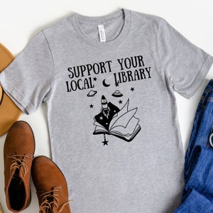 May include: Gray t-shirt with the text "Support Your Local Library" and a black and white illustration of a book with a rocket, stars, and a crescent moon.