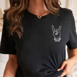 May include: Black t-shirt with a white outline of a hand making the rock and roll hand gesture. The hand is wearing a bracelet with spikes.