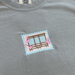 May include: A light gray t-shirt with a small embroidered design of a window with pink flowers. The t-shirt has a tag that says "Comfort Colors".