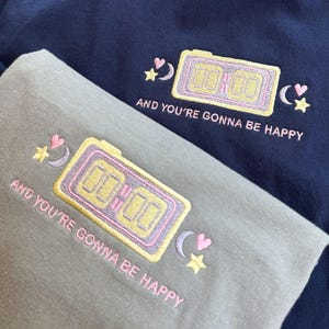 Puede incluir: Dos sudaderas, una azul marino y otra gris, con diseños bordados. Cada una presenta un reloj digital, estrellas y lunas crecientes, con el texto "AND YOU'RE GONNA BE HAPPY" en rosa. La etiqueta Comfort Colors es visible en la camisa azul marino.